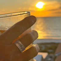 Hand holding a drink at sunset, showcasing the Lupine Adjustable Ring with simple silhouette of a lupine flower.