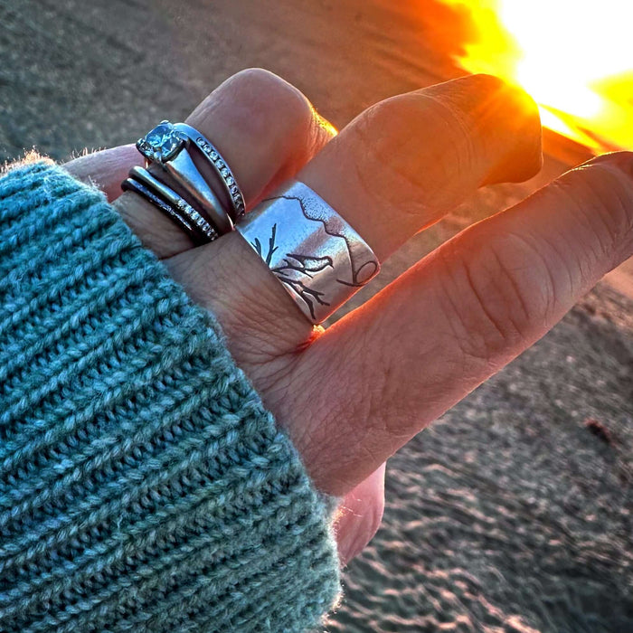 Hand wearing Mtn Bluebird Adjustable Ring with mountainscape design at sunset, showcasing layered rings.