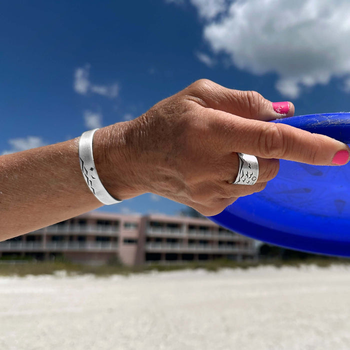 Hand wearing Fly Over cuff bracelet, showcasing nature design on the beach with blue frisbee.