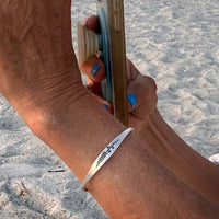 A woman wearing a delicate Lupine Flower Cuff Bracelet while holding a phone on a sandy beach.