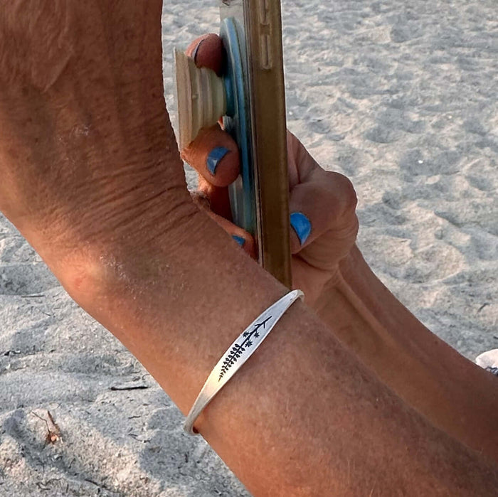 A woman wearing a delicate Lupine Flower Cuff Bracelet while holding a phone on a sandy beach.