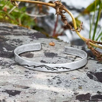 Mountainscape Mountain Cuff Bracelet displayed on a rock, showcasing its detailed mountain design and silver finish.