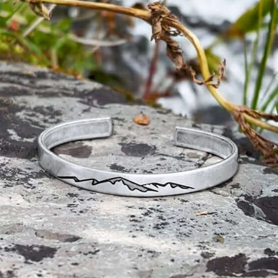 Mountainscape Mountain Cuff Bracelet displayed on a rock, showcasing its detailed mountain design and silver finish.
