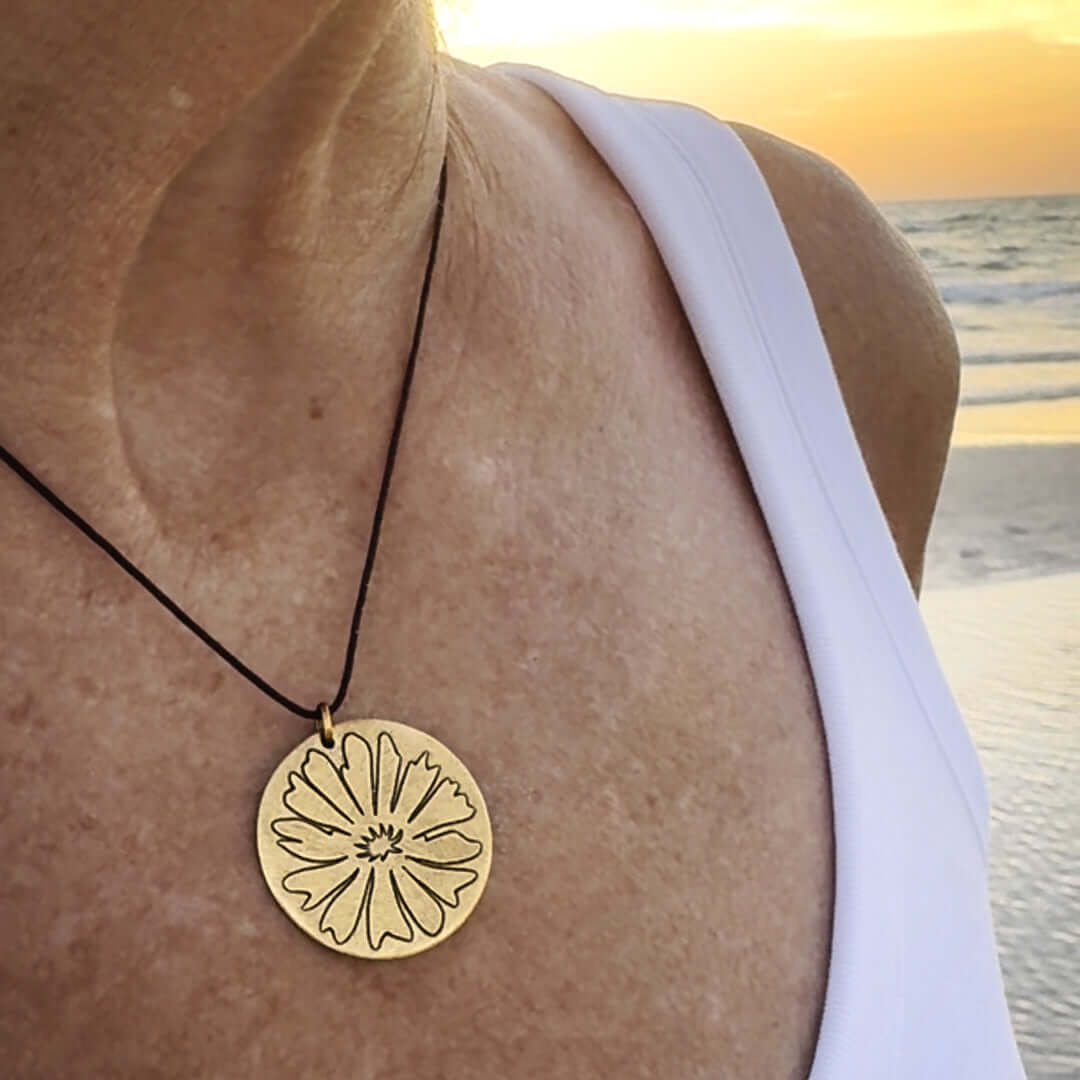 Close-up of a woman wearing a Bitterroot Necklace with a gold pendant featuring a flower design at sunset by the beach.