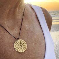 Close-up of a woman wearing a Bitterroot Necklace with a gold pendant featuring a flower design at sunset by the beach.