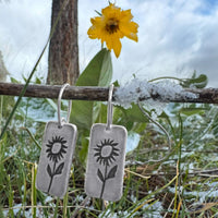 Balsamroot flower earrings in matte silver, displayed with a yellow flower and greenery in a natural setting.
