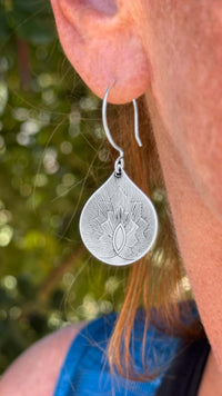 Close-up of delicate silver Petal Earrings, showcasing a floral design and teardrop shape, worn by a person outdoors.