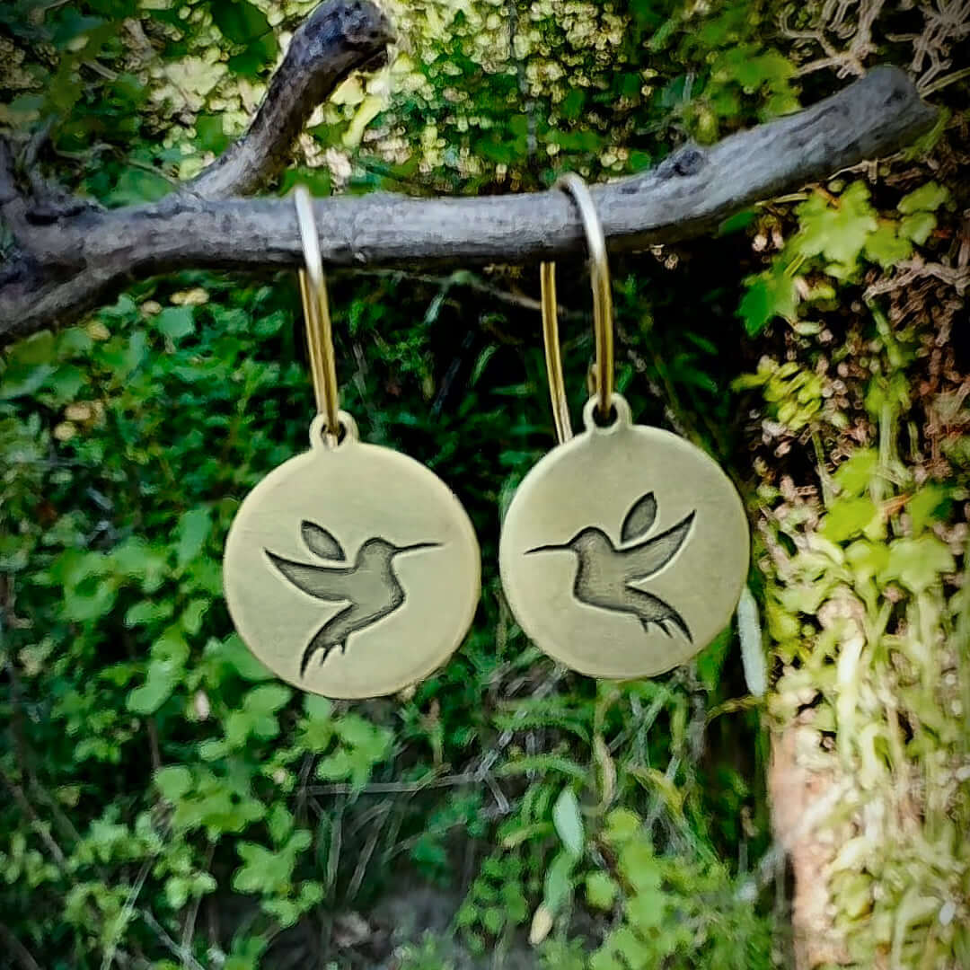 Antique brass hummingbird earrings with engraved design, elegantly displayed on a branch amidst greenery.