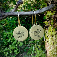 Antique brass hummingbird earrings with engraved design, elegantly displayed on a branch amidst greenery.