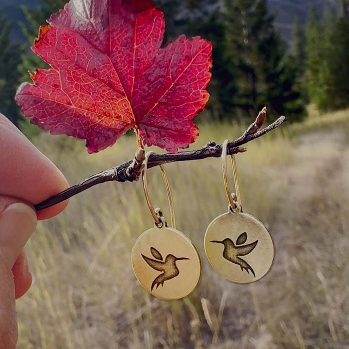 Hummingbird earrings hanging from a twig, surrounded by fall foliage in a serene outdoor setting.