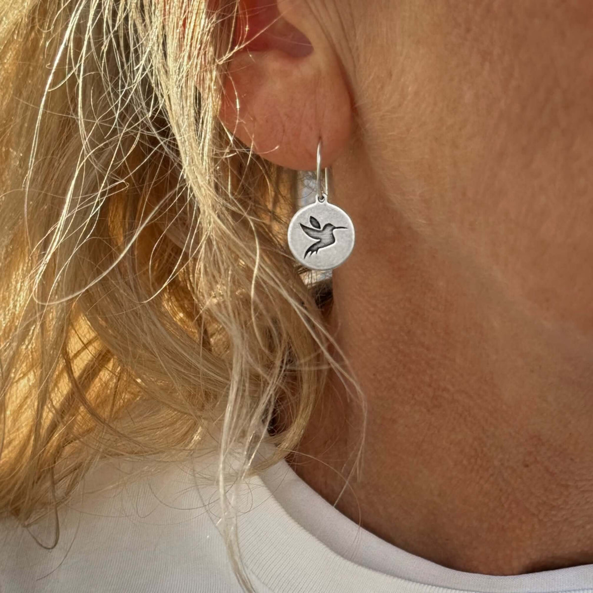 Close-up of sterling silver hummingbird earrings worn by a person with blonde hair.