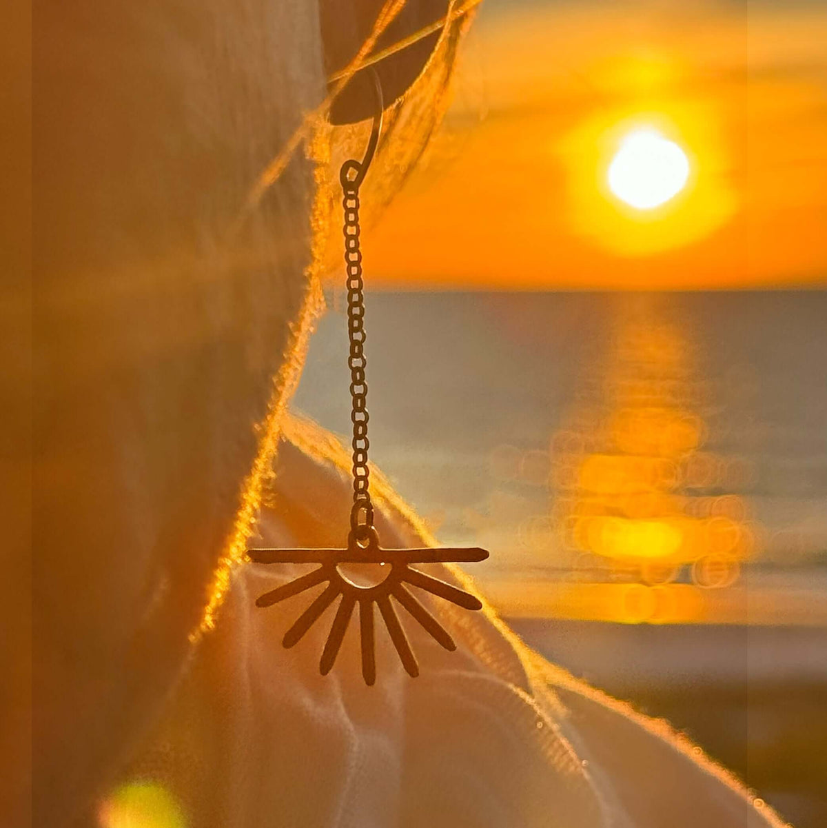 Sunbeam Earrings featuring delicate sunray design, swinging on a rolo chain against a sunset backdrop.
