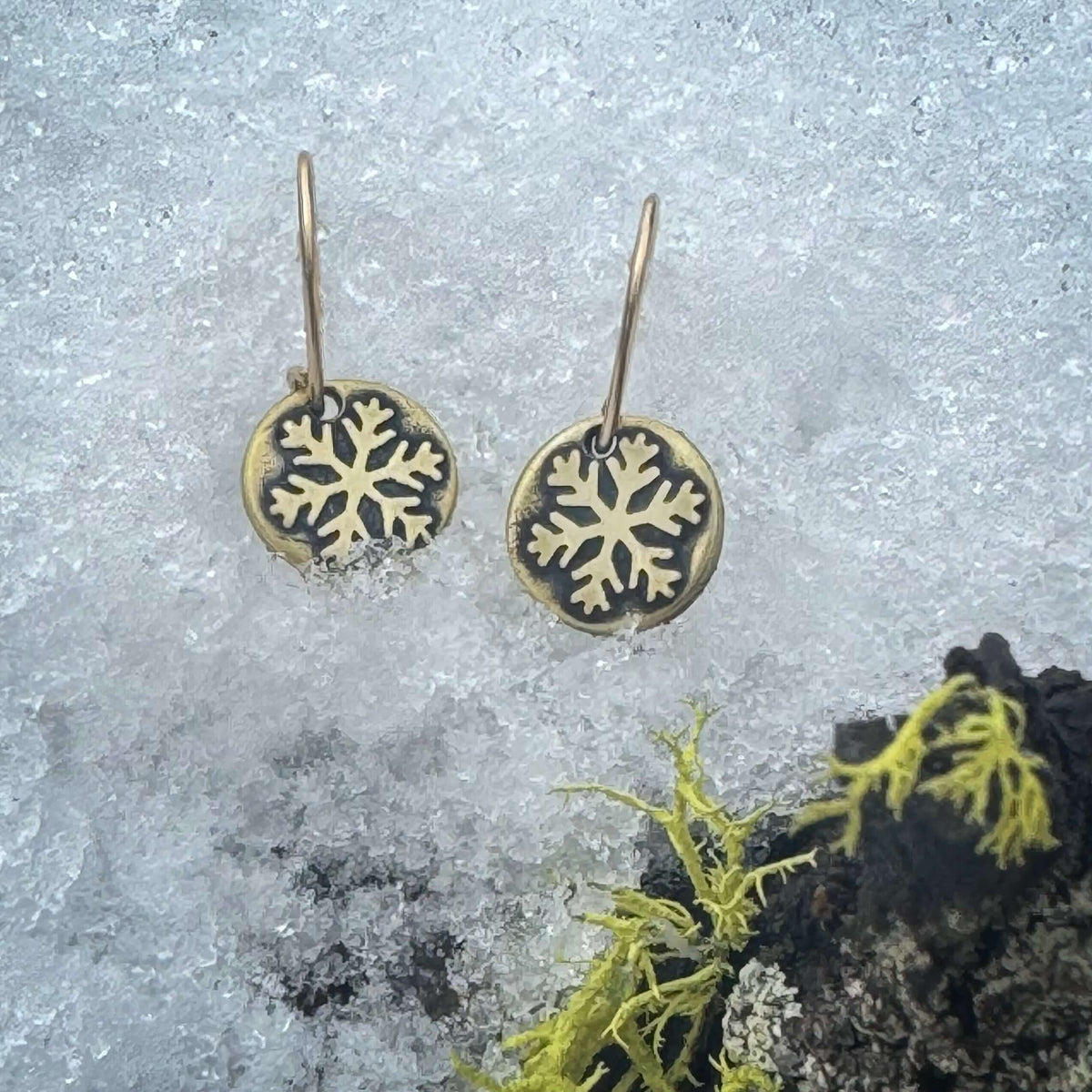 Powder Snowflake Earrings hanging on snow with moss in the background, featuring an antique brass finish.