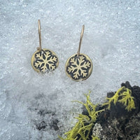 Powder Snowflake Earrings hanging on snow with moss in the background, featuring an antique brass finish.