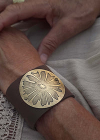 A close-up of a wrist adorned with the Wildflower Leather Cuff, a wide brown bracelet featuring a large round antique brass floral design, as hands rest on light-colored textured fabric.