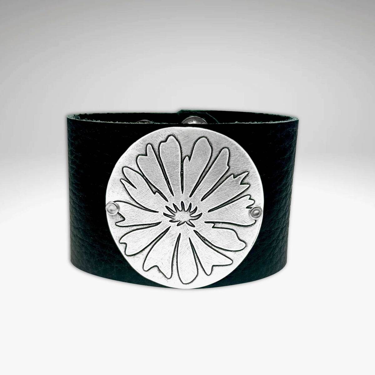 The Wildflower Leather Cuff is a black leather bracelet with a prominent round matte silver plate on the front, engraved with an abstract flower design. The background features a subtle gradient from gray to white.