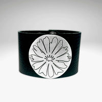 The Wildflower Leather Cuff is a black leather bracelet with a prominent round matte silver plate on the front, engraved with an abstract flower design. The background features a subtle gradient from gray to white.