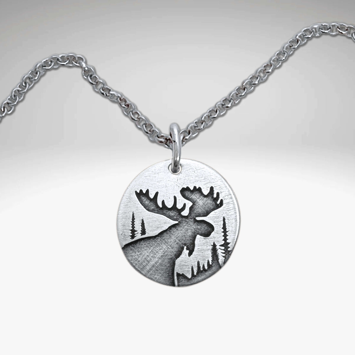 The Creek Moose Necklace showcases a matte silver pendant with an engraved moose silhouette and forest scene on a round medallion.