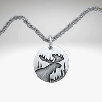 The Creek Moose Necklace showcases a matte silver pendant with an engraved moose silhouette and forest scene on a round medallion.