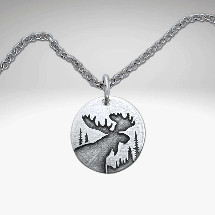 The Creek Moose Necklace showcases a matte silver pendant with an engraved moose silhouette and forest scene on a round medallion.