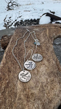 Creek Moose Earrings showcasing detailed moose designs and necklace in matte silver finish on a rustic wooden background.