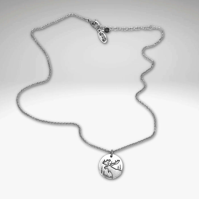 The Creek Moose Necklace features a matte silver round pendant engraved with a moose and pine trees on an 18" chain.