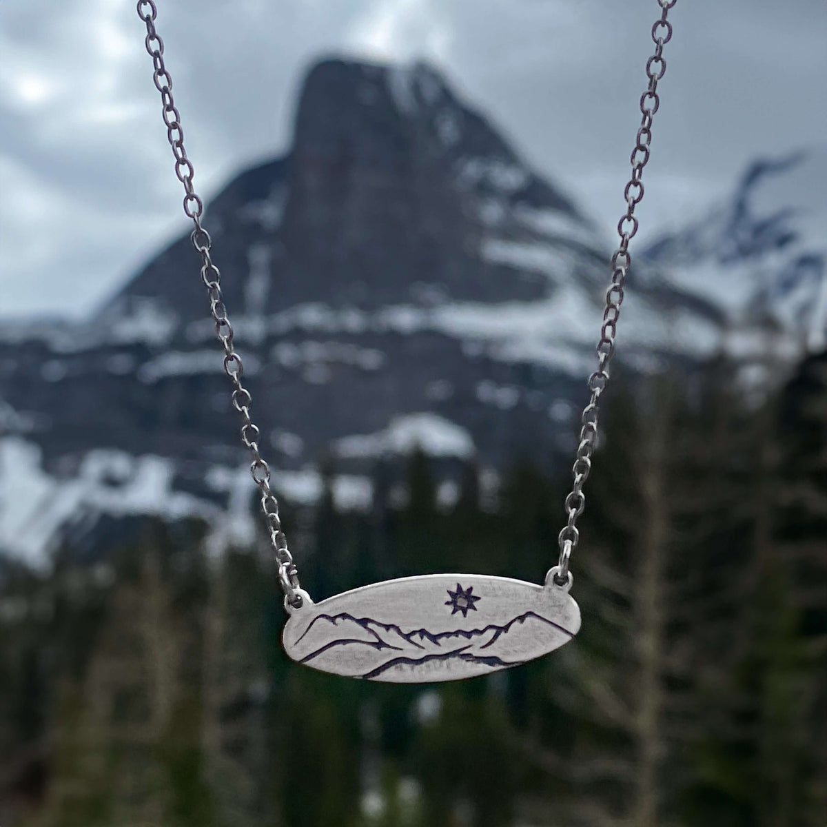 Glacier View Necklace featuring mountain design on a matte silver pendant with chain against a mountainous backdrop.