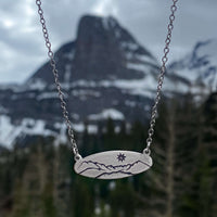 Glacier View Necklace featuring mountain design on a matte silver pendant with chain against a mountainous backdrop.