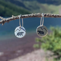 Mountain Bear Earrings showcased on a branch with a natural background, featuring etched mountain and bear design in matte silver.