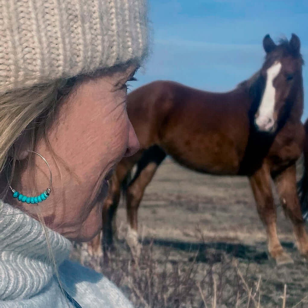 A woman wearing Shun Howlite Beaded Hoop Earrings with turquoise howlite beads, smiling next to a horse in a field.