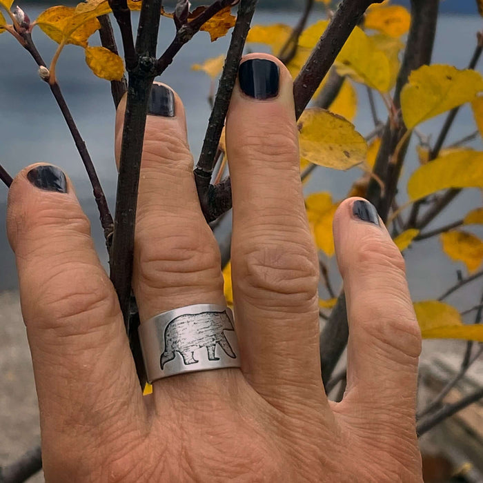 Bear Boy Adjustable Ring worn on a hand with dark nails, surrounded by autumn leaves near water.