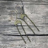Moonfall Earrings in antique brass with dangly chains on rustic wooden background.