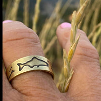 Fishing Adjustable Ring featuring a fish design etched in antique brass, worn on a hand in a grassy outdoor setting.