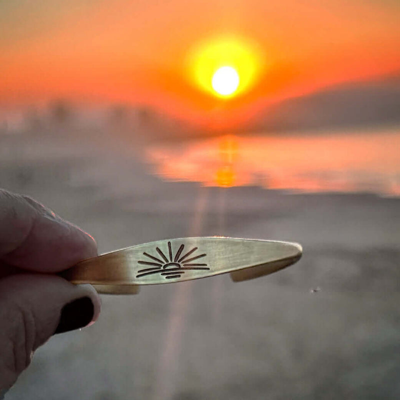 A hand holds a brass bracelet with a sunrise design, with a blurred beach and vibrant orange sunset in the background over the water.