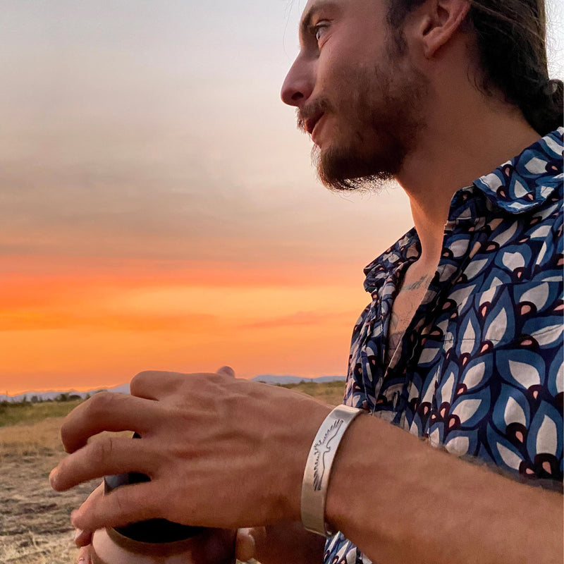 A man with a patterned shirt and a silver bracelet holds a cup outdoors at sunset, with a colorful sky and landscape in the background.