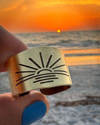 Hand holding a Sunrise Adjustable Ring with sun design against a sunset beach background.