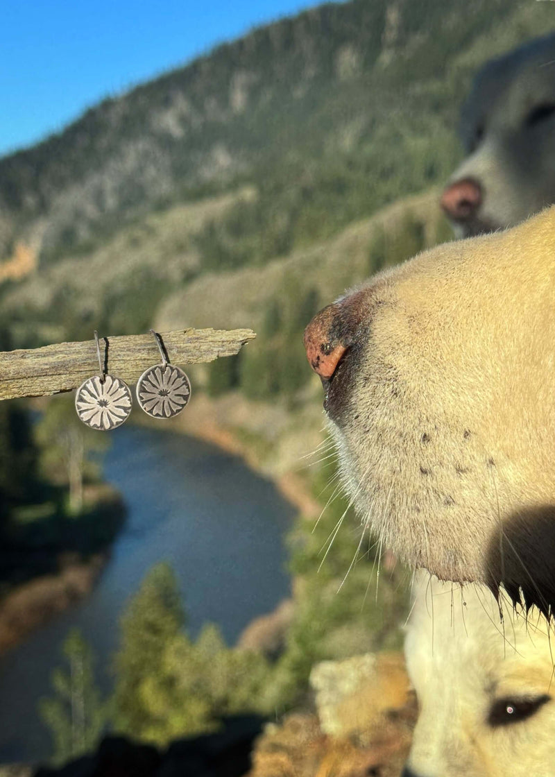 Bitterroot Flower Earrings displayed on a branch with a dog in the foreground near a river and mountains.