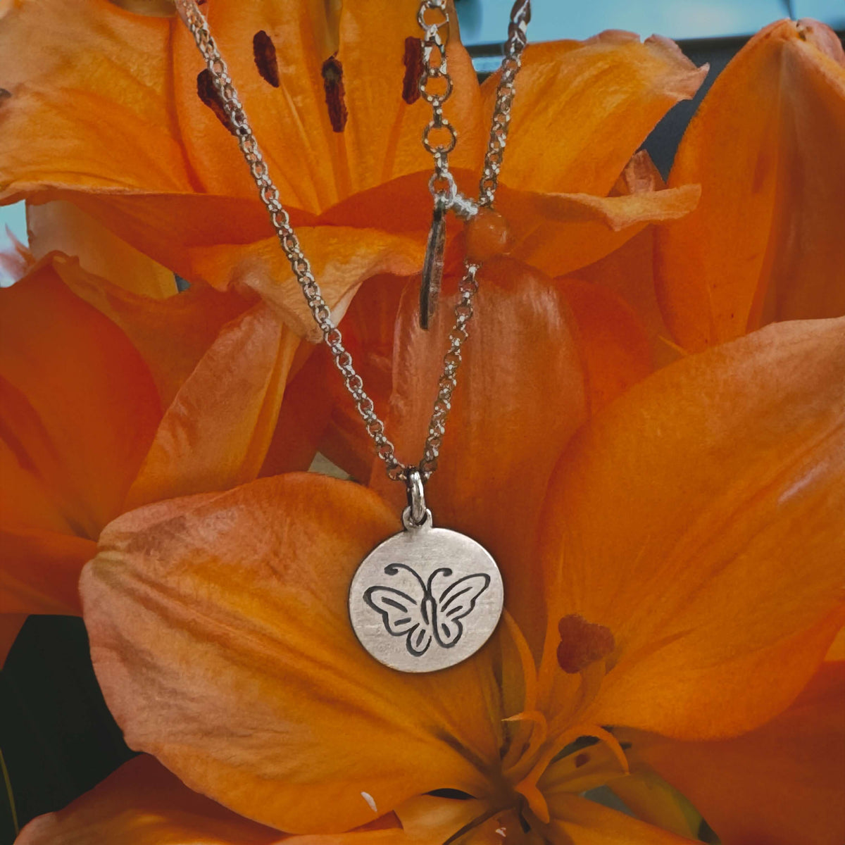 Butterfly Necklace displayed on vibrant orange flowers, showcasing its etched butterfly design and adjustable chain.