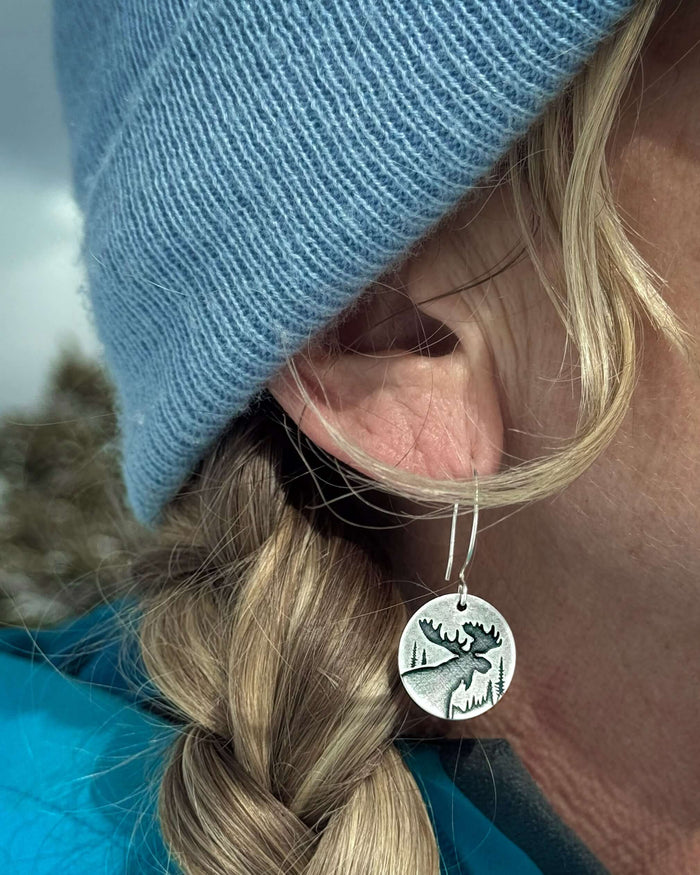 Creek Moose Earrings featuring a moose design on a silver circular charm worn by a model outdoors.