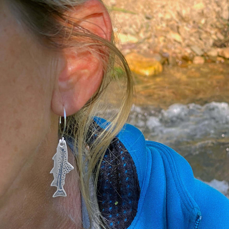 Daphne Lorna Fishing Trout Earrings – Handmade Montana Wildlife Jewelry, Matte Silver or Antique Brass worn by a woman near a river.