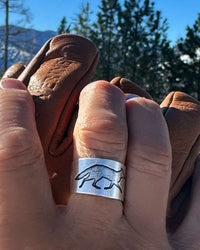 Woman's finger wearing Fox Run Adjustable Ring in Matte Silver, featuring a running fox design, against a natural outdoor backdrop.