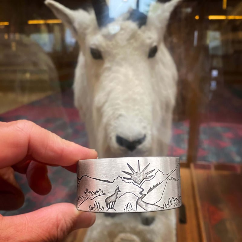 Going to the Sun Road Cuff Bracelet featuring mountains, a sun goat, and road design in front of a mountain goat display.