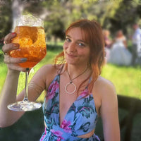 Woman enjoying a drink while wearing a Hula Necklace with a circle design, in a floral dress at an outdoor event.