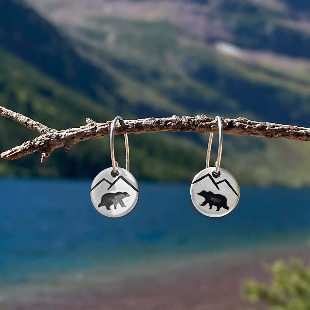 Mountain Bear Earrings hanging on a twig against a scenic lake background, showcasing bear and mountain designs.