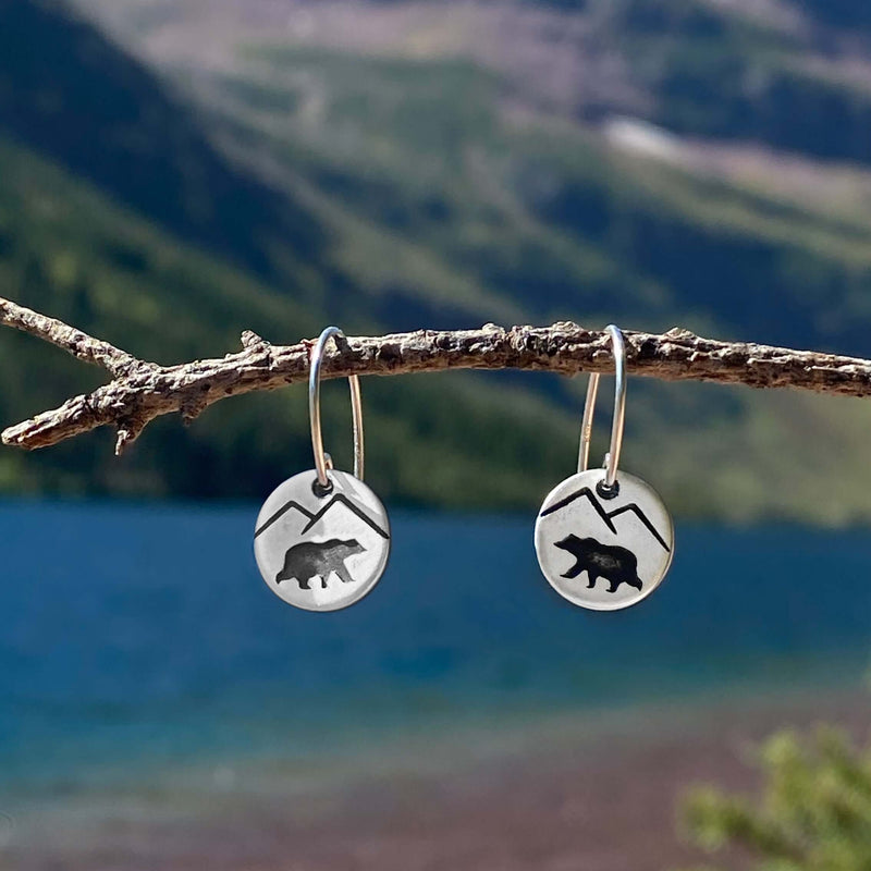 Mountain Bear Earrings hanging on a twig against a scenic lake background, showcasing bear and mountain designs.