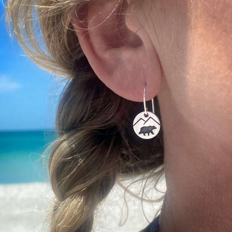 Person wearing Mountain Bear Earrings with a bear and mountain design on a beach background.