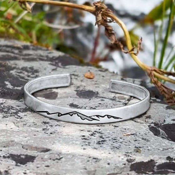 Mountainscape Mountain Cuff Bracelet displayed on a rock, featuring a mountain design etched on a matte silver finish.