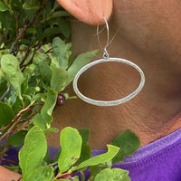 Open Space Earrings featuring a lightly hammered open ellipse design worn by a person surrounded by greenery.