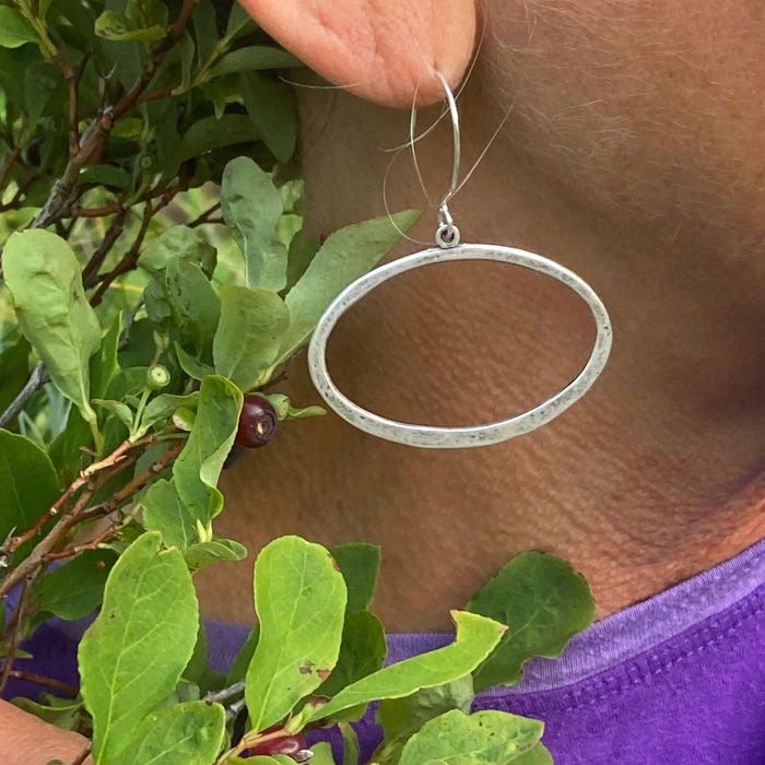 Open Space Earrings featuring a lightly hammered open ellipse design worn by a person surrounded by greenery.