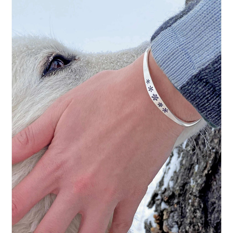A close-up of a hand wearing the Powder Signet Cuff Bracelet adorned with snowflakes next to a snowy background.